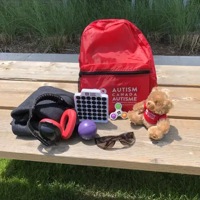 Contents of Autism Sensory Kit on a picnic table - includes a red backpack, teddy bear, sunglasses, headphone, fidget spinner and a blanket