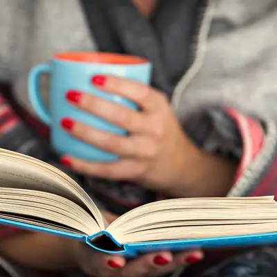 Woman Wrapped In A Blanket Reading A Book And Holding A Mug
